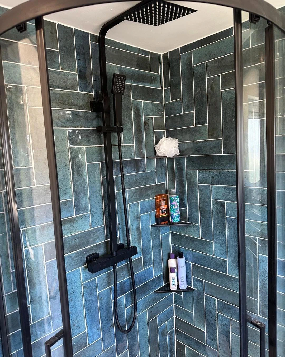 Teal tiled shower enclosure with black rainfall shower and corner storage shelves