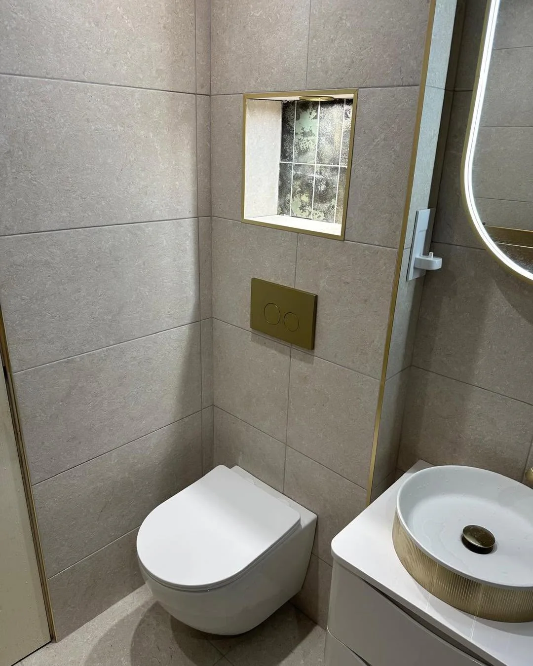 Wall-hung toilet, bronze flush plate and recessed shelf within pale stone tiles