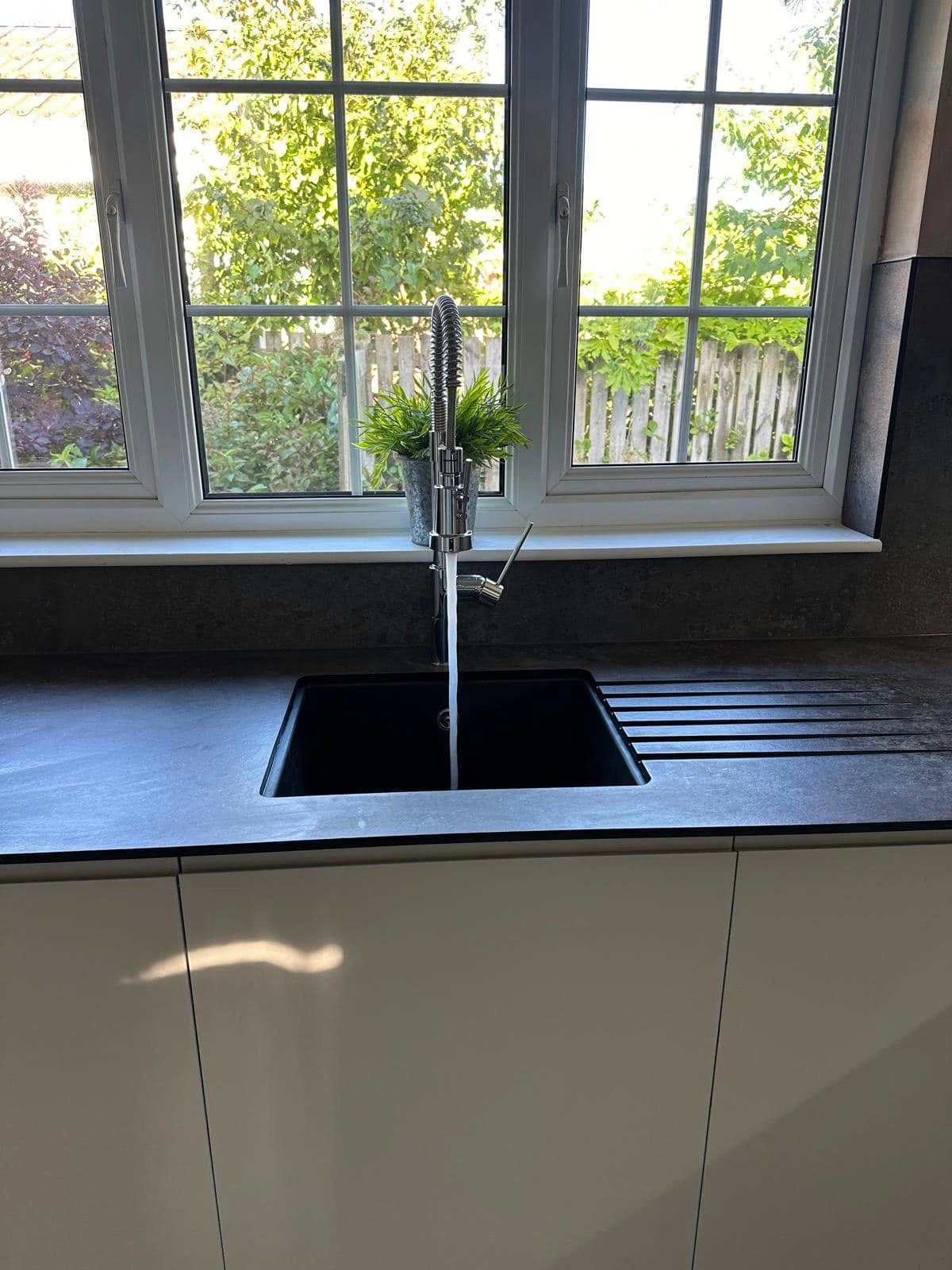 Black kitchen sink and spring-neck tap set beneath wide garden-facing windows