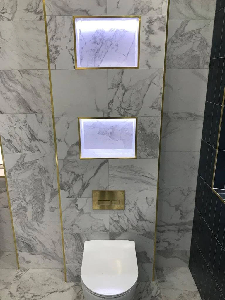 Wall-hung toilet beneath two illuminated recessed shelves with brass trim