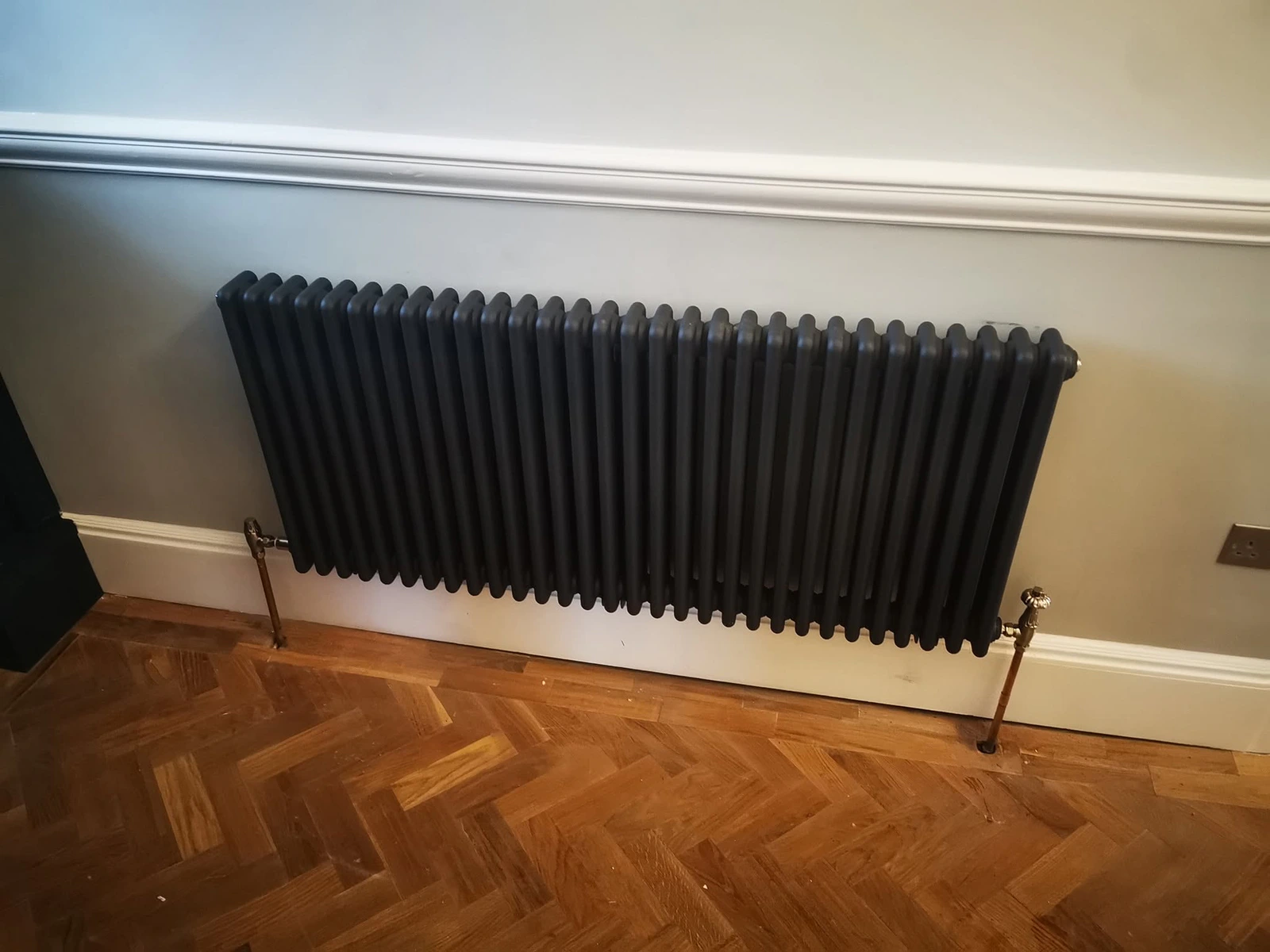 Dark traditional radiator with exposed pipework above herringbone timber flooring