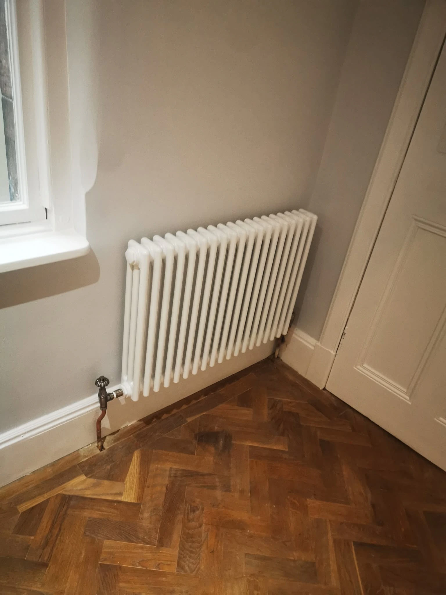 White traditional radiator fitted beside a window above herringbone timber flooring